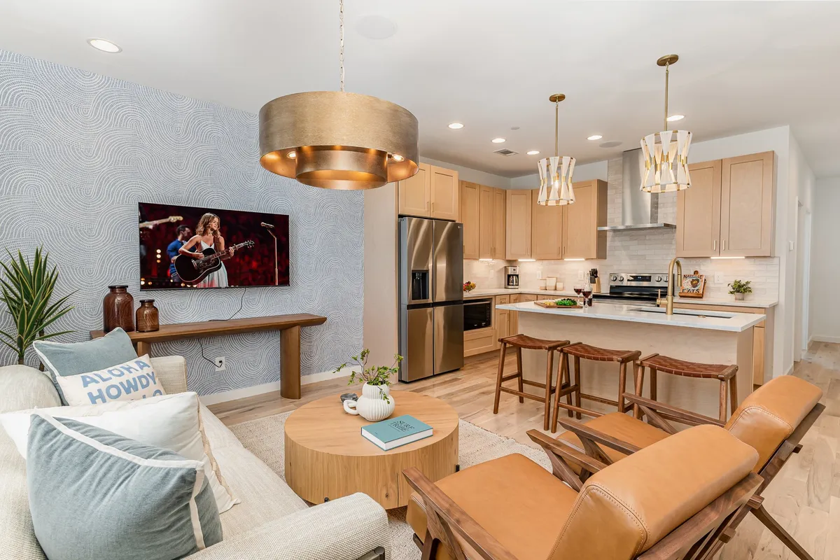 The image showcases a bright, open-concept living area with a cozy sofa adorned with decorative pillows, a rustic wooden coffee table, a modern kitchen island with stylish bar stools, and a wall-mounted TV displaying a musician—inviting guests to gather, relax, and enjoy entertainment together. Elegant pendant lighting completes the scene, adding warmth and flair.