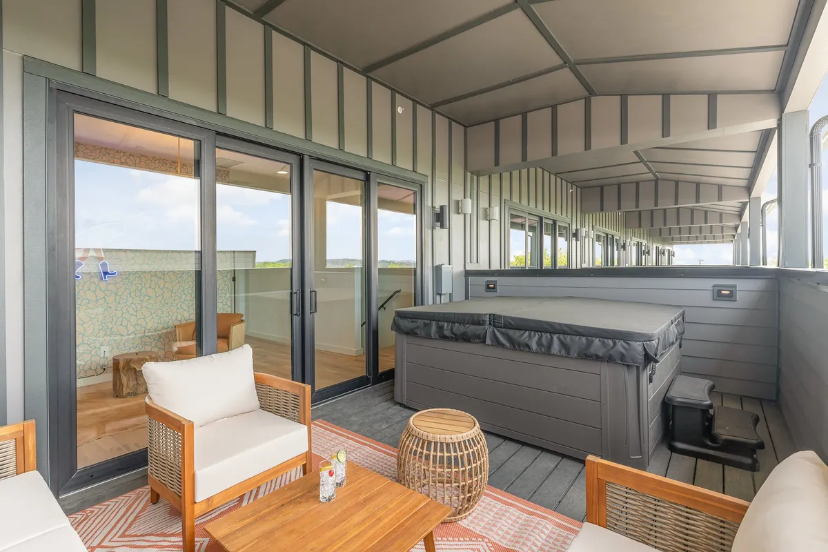 A spacious outdoor deck featuring a bubbling hot tub, comfortable lounge seating, a wooden table, and a decorative rug. The scene is bathed in sunset light, suggesting a relaxed, inviting atmosphere ideal for socializing or unwinding. The deck is elevated, offering views and a private retreat.