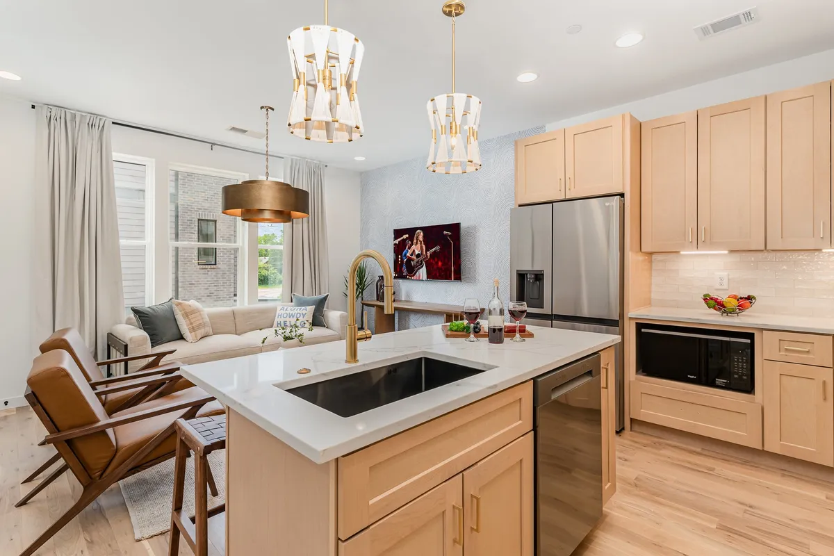 This image displays an open-concept interior space with a kitchen island featuring gold pendant lighting, a stylish seating area with a comfortable couch, and a television mounted on the wall. The space feels spacious, modern, and ideal for both relaxing and gathering with friends or family.