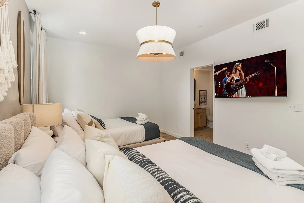 A modern bedroom with two beds adorned with decorative pillows, a wall-mounted smart TV, a chandelier light fixture, and a stylish wall hanging. The mood is comfortable and relaxed, ideal for unwinding or enjoying a movie night.