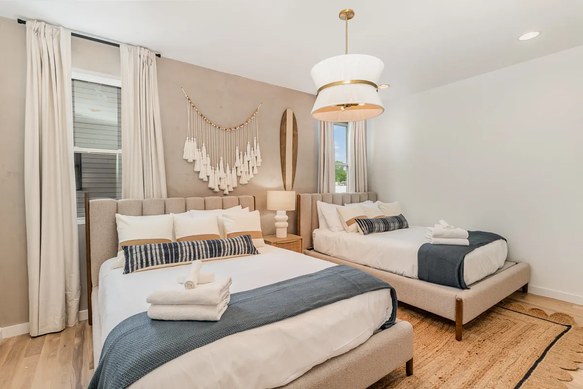 A beautifully appointed bedroom with two large beds, a decorative wall hanging, a stylish light fixture, and a soft, textured rug. The room is bathed in gentle natural light, creating a relaxed and tranquil atmosphere.