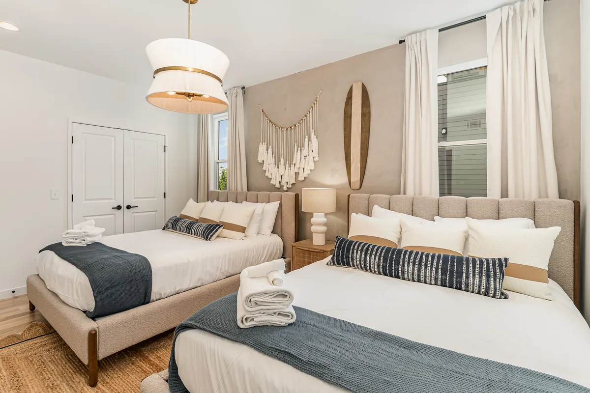 This image features a serene bedroom with two beds, accented by decorative wall art and a pendant light fixture. Natural light streams through a window, illuminating the calm and inviting space. The decor is elegant yet cozy, emphasizing comfort and relaxation.