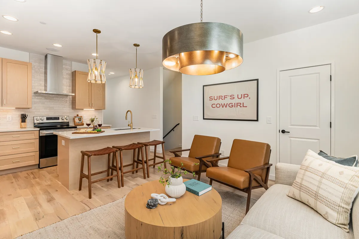 The photo highlights a bright, inviting kitchen with a large island, contemporary light fixtures, wall artwork, and a welcoming seating arrangement. The setup encourages group entertaining, shared meals, and lively conversation around the kitchen counter.